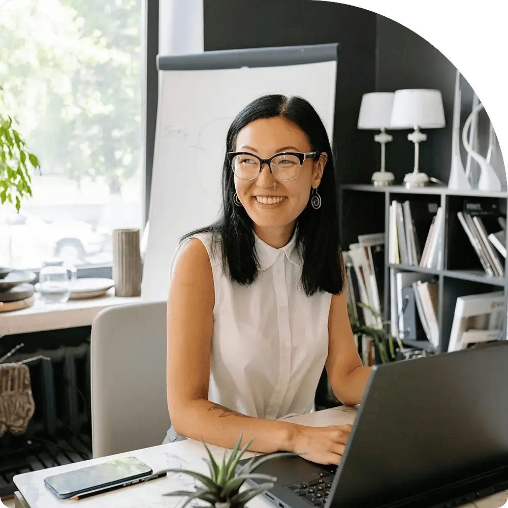 A bright, inviting office scene: a smiling person with straight, shoulder-length black hair and black-framed glasses sits at a desk, hands poised over a laptop.