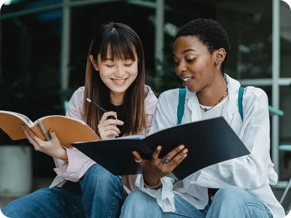 Two students sit outdoors, smiling and studying together. One holds a notebook, the other a folder.