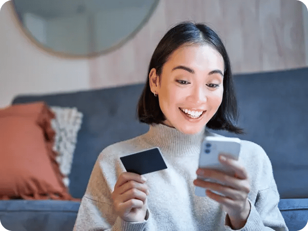 A woman sits on the floor, smiling as she holds a smartphone and a credit card.