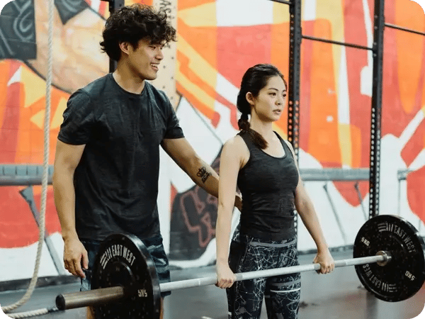 Man coaches women in gym. She is holding a large weight. Both wear workout gear.