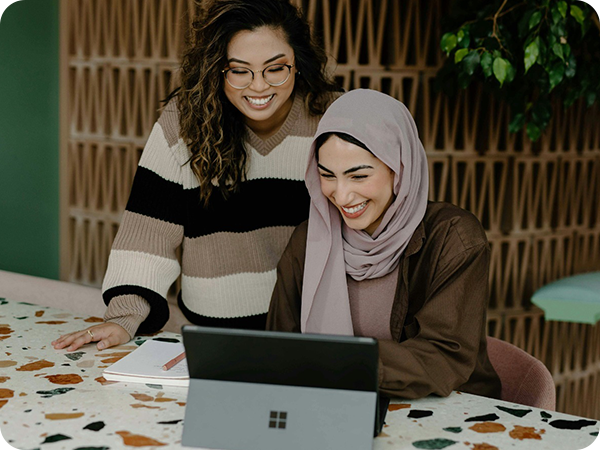 Two women smiling as they look at a laptop. One wears glasses and a striped sweater, the other a hijab. The setting is a modern, stylish room.