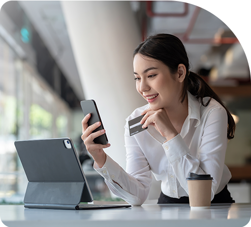 A woman in a white shirt smiles while holding a credit card and smartphone. She sits at a table with a tablet and coffee cup in a modern, bright setting.
