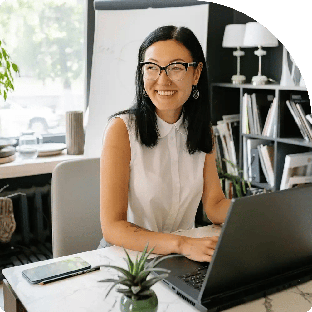 A bright, inviting office scene: a smiling person with straight, shoulder-length black hair and black-framed glasses sits at a desk, hands poised over a laptop.