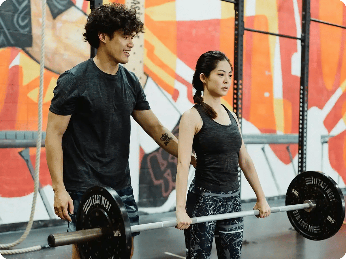 Man coaches women in gym. She is holding a large weight. Both wear workout gear.