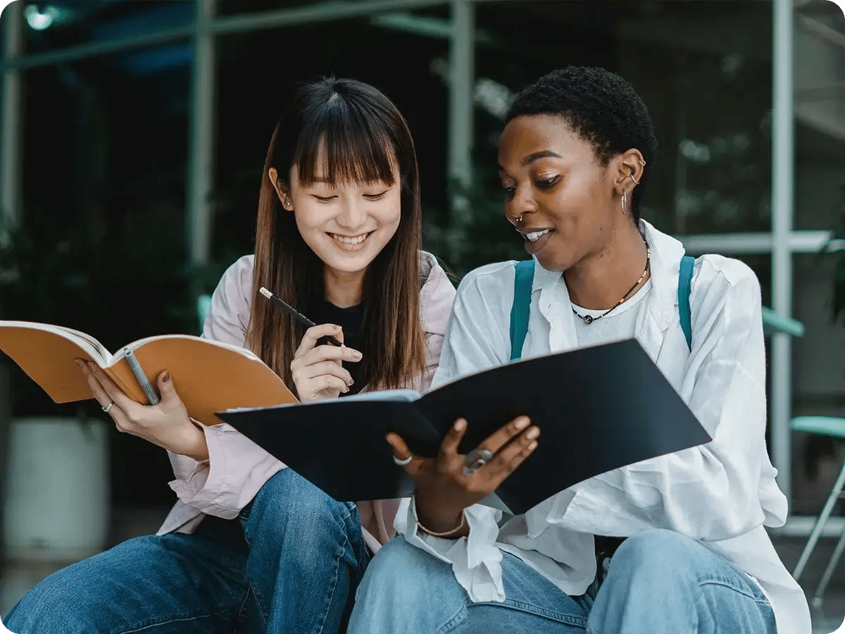 Two young women sitting together, one holding a notebook and a pen, while the other has an open folder. They appear to be studying and smiling in a casual indoor setting.