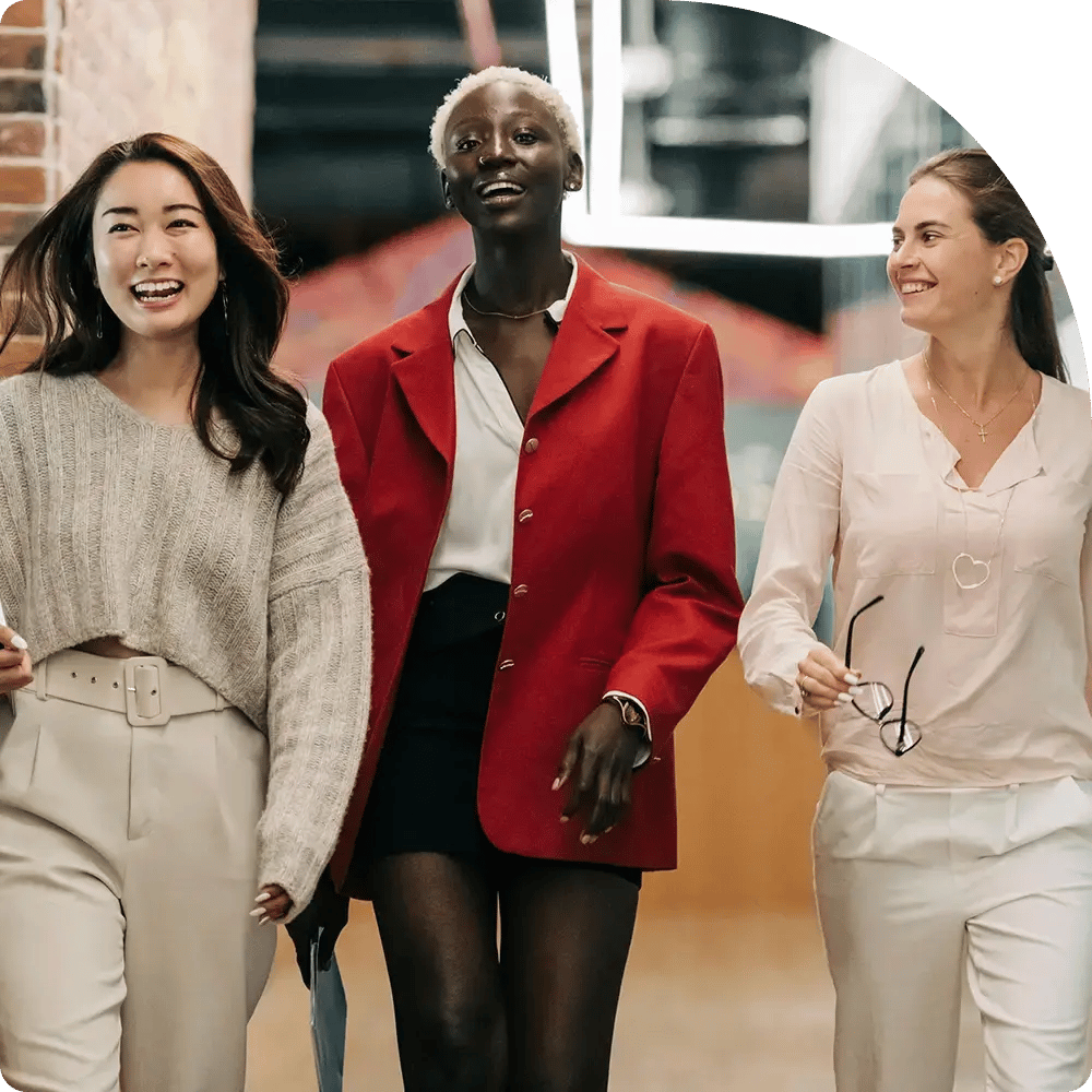 Three women walking confidently indoors, smiling. One wears a gray sweater, another a red blazer, and the third a white blouse. Bright, positive atmosphere.