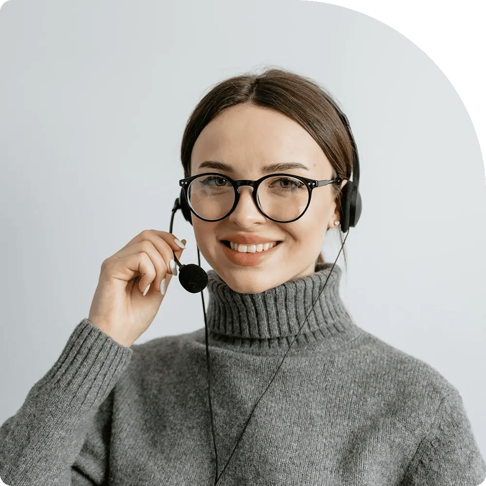 A smiling woman wearing glasses and a headset, with a hand on her microphone, against a light background.