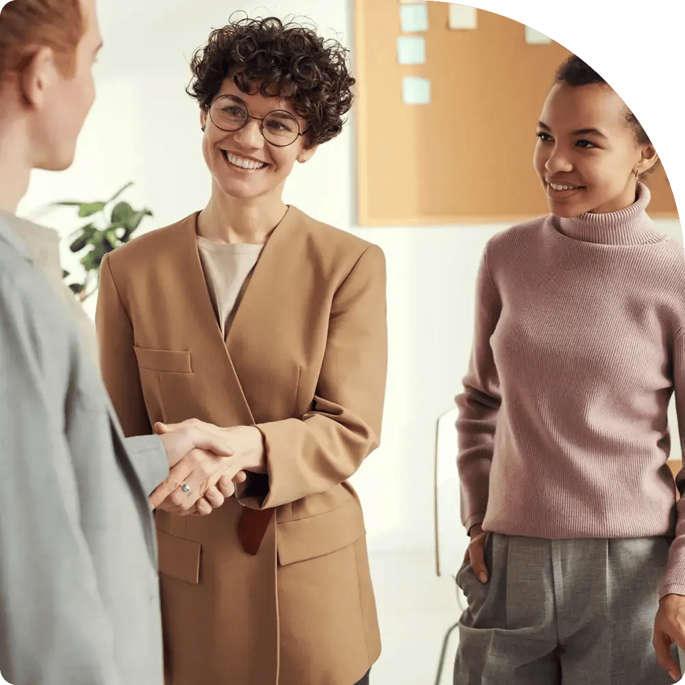 Two women smiling warmly during a handshake. One wears glasses and a tan blazer, the other wears a pink sweater, conveying a positive, professional meeting.