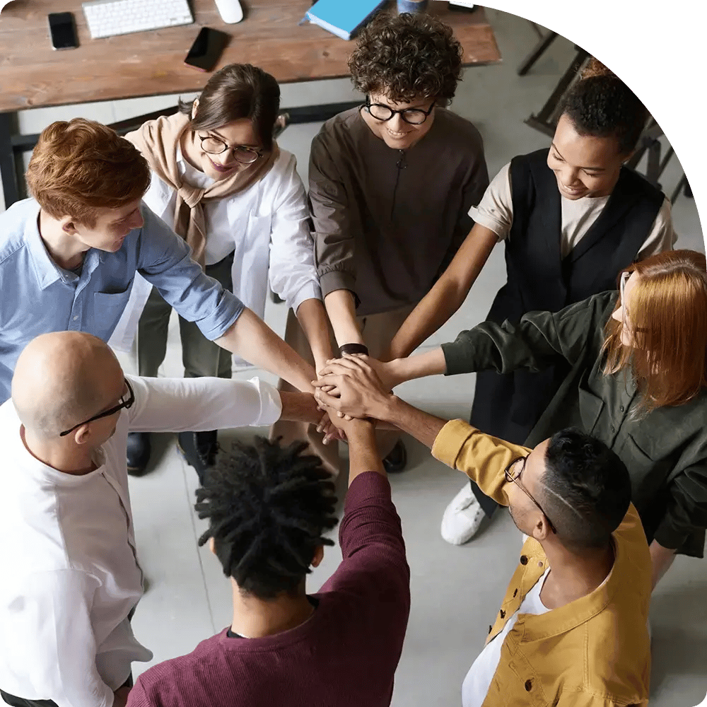 Diverse group of eight people stand in a circle, hands joined in the center. The atmosphere is collaborative and supportive, conveying teamwork.