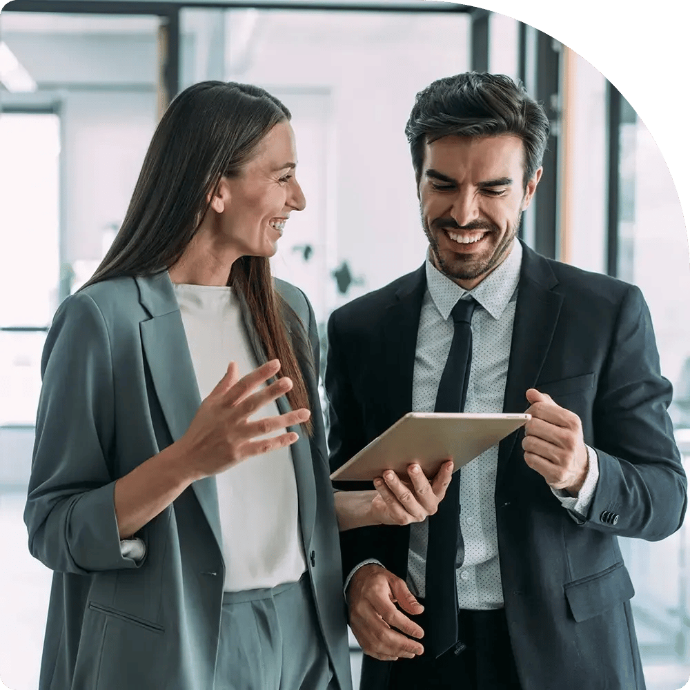 A woman and a man in formal business attire smile while looking at a tablet.