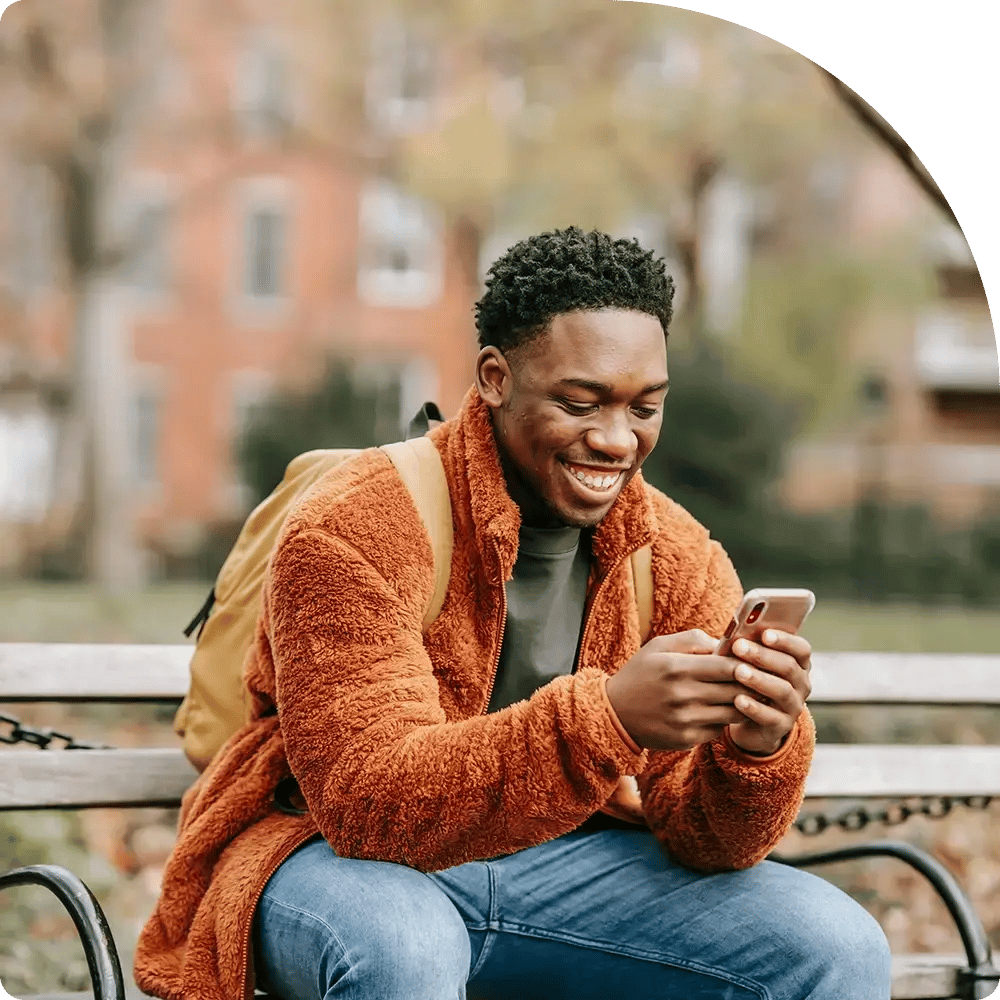 A person in an orange fleece jacket sits on a park bench, smiling at their phone.
