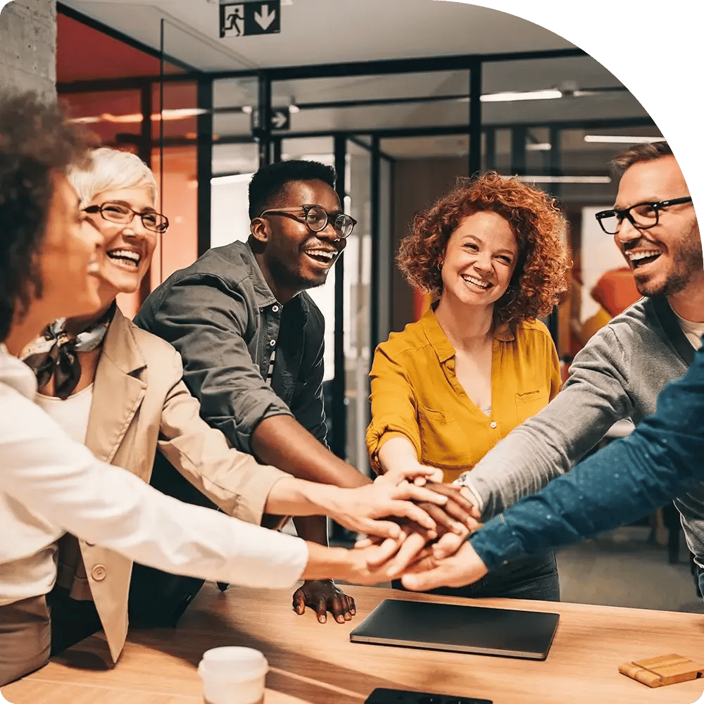 A diverse group of six people smiling and placing their hands together in a cheerful office setting, symbolizing teamwork and unity.