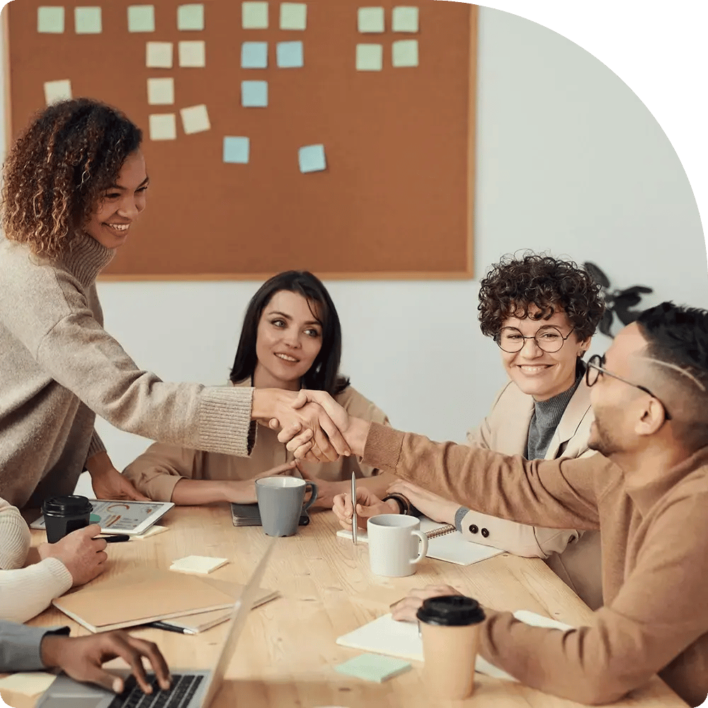 A diverse group of four people in cozy sweaters sit around a wooden table, smiling, as two of them shake hands. Colorful sticky notes on a corkboard.