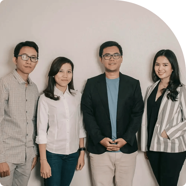 A group of four people smiling, standing in a row against a plain background. They are dressed in casual to business casual attire, conveying a friendly and professional tone.