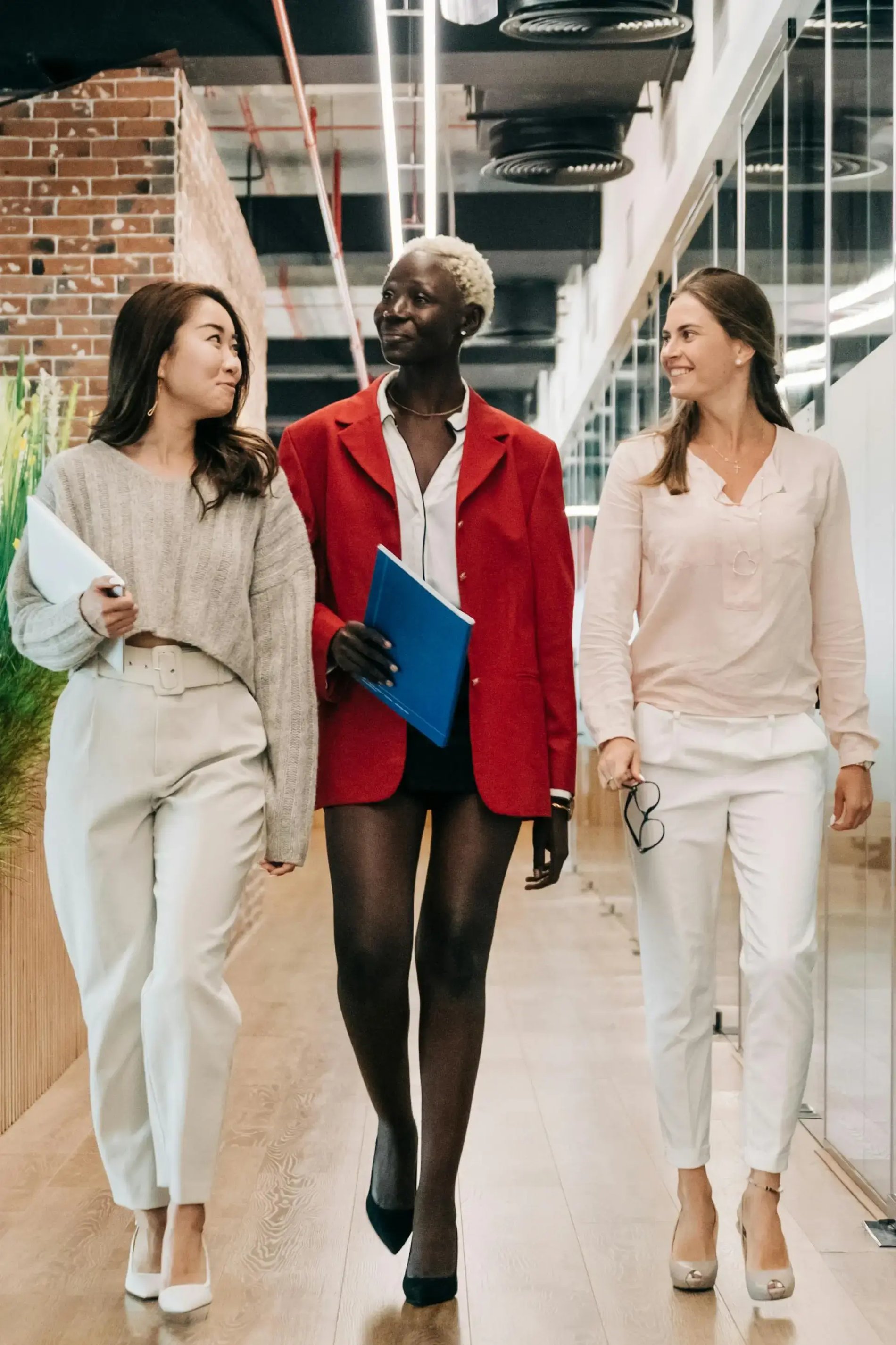 Three women walk together, smiling and chatting. The woman on the left holds a laptop, the center wears a red blazer, and the right holds glasses.