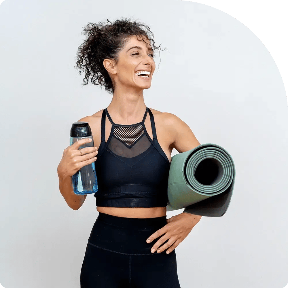 Woman smiling, holding a yoga mat and water bottle, wearing a black sports bra and leggings against a plain background. She appears cheerful and energized.