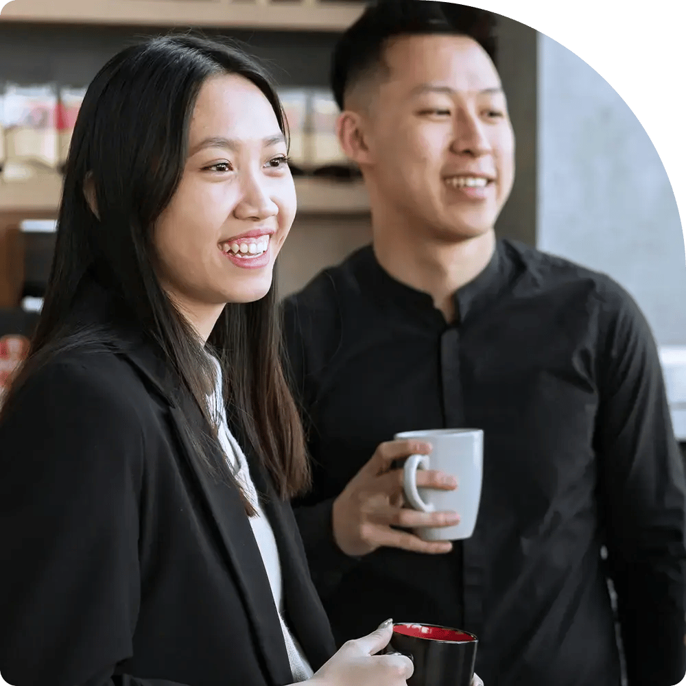 Two people smile and hold mugs in a relaxed setting. They are dressed in black, enhancing the professional tone.