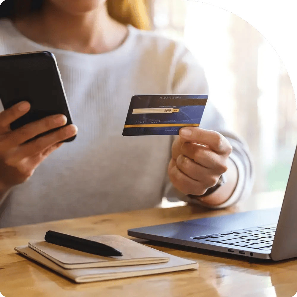 A person holding a credit card and smartphone above a notebook and pen on a wooden table, with a laptop nearby, conveying online payments.