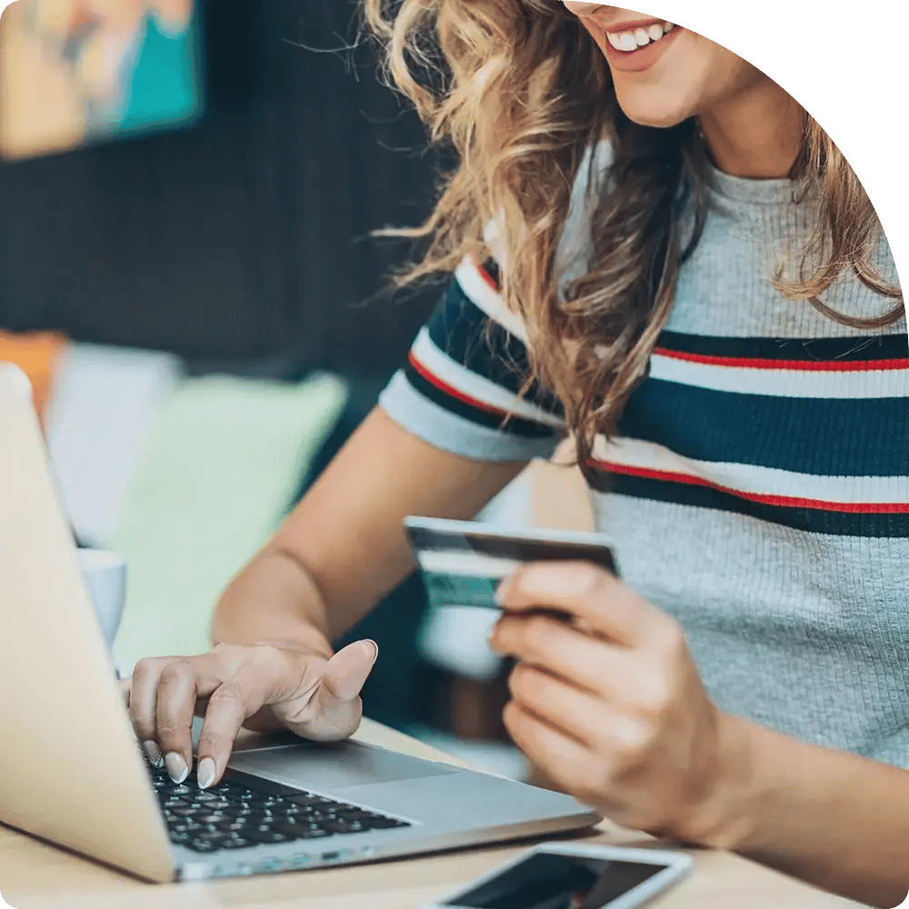 A woman with wavy hair smiles while holding a credit card and typing on a laptop.