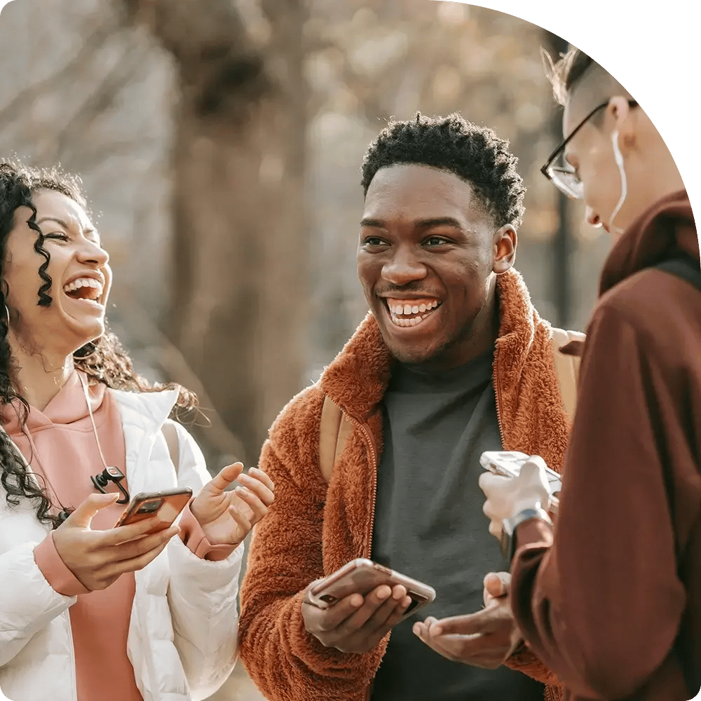 A group of three young adults at outdoors, laughing and holding smartphones.