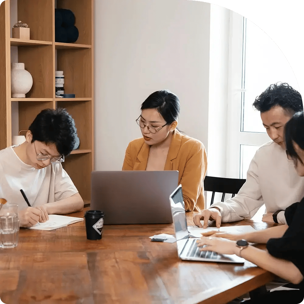 A diverse group of four people engages in a focused meeting at a wooden table, with laptops and papers. Shelves with decor are in the background. Light filters through a window, creating a collaborative, productive atmosphere.