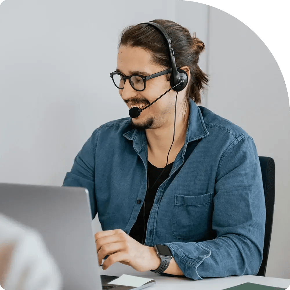 A smiling person with glasses and a headset works at a laptop. They wear a blue shirt and smartwatch, creating a focused yet relaxed atmosphere.