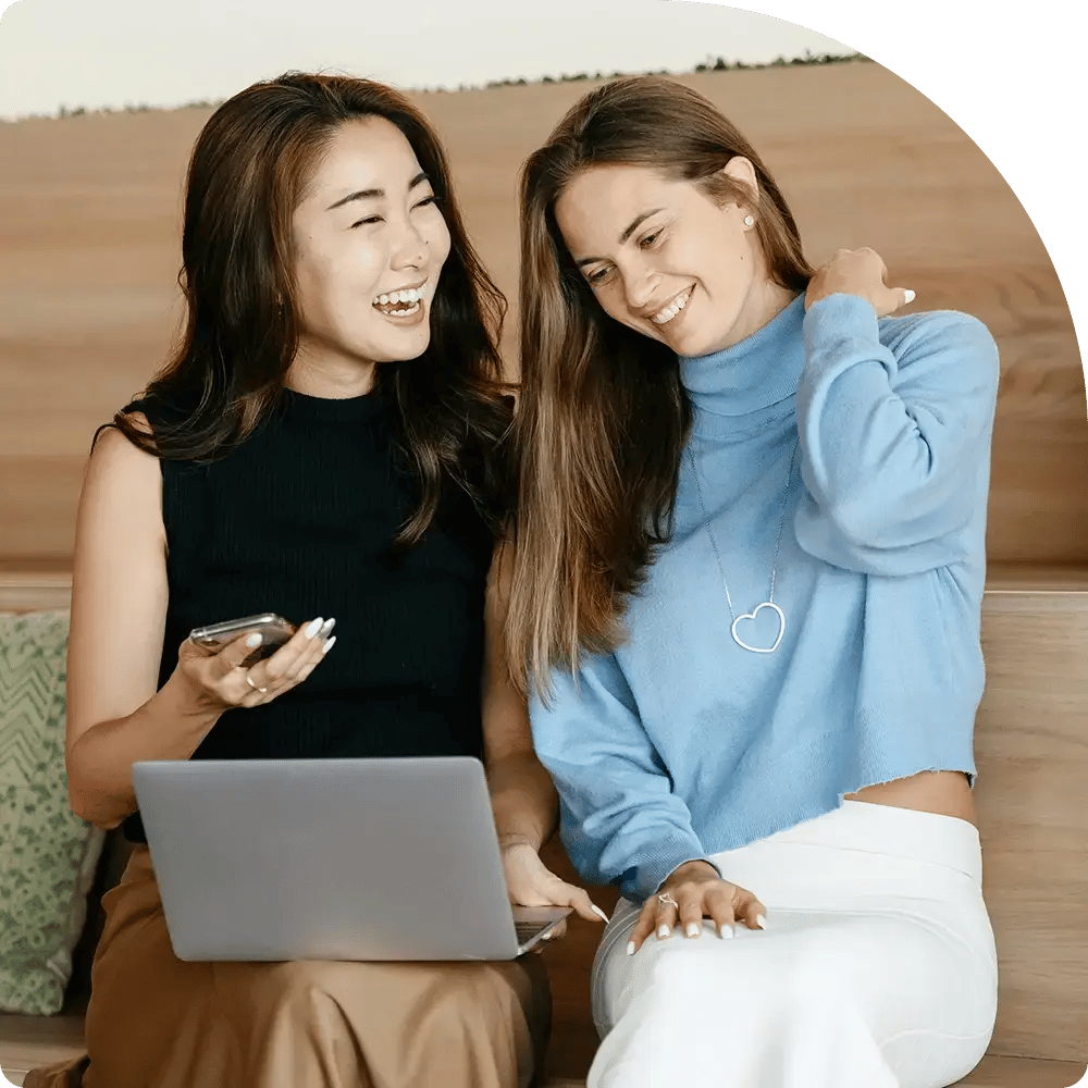 Two women sitting on a sofa, looking at a laptop and smiling.