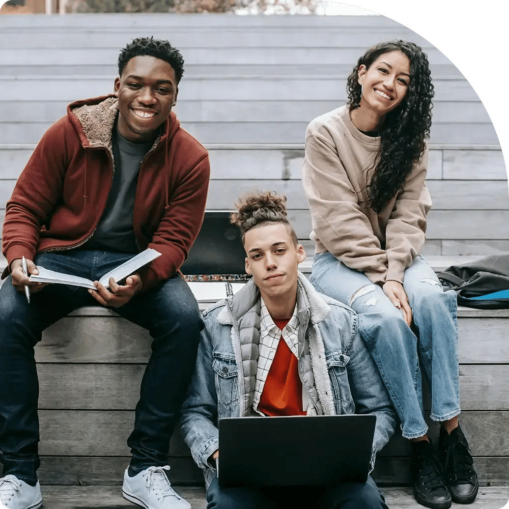 A group of three young adults sitting on steps, smiling at the camera. One person is working on a laptop, while another holds a device.