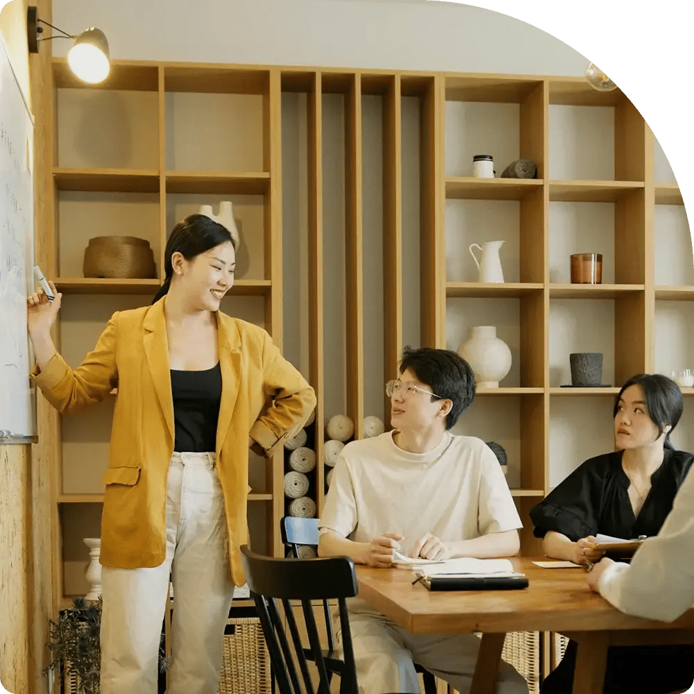 Woman in a yellow blazer talks to a seated man in a cozy study with a wooden shelf.