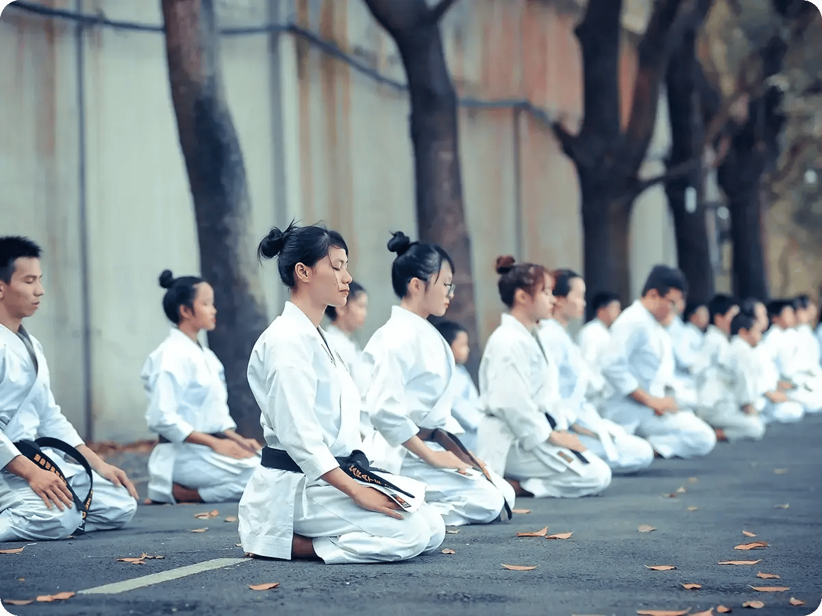 Practitioners in white karate uniforms kneel in meditation on an outdoor path, lined with trees. Their focused expressions convey calm and discipline.