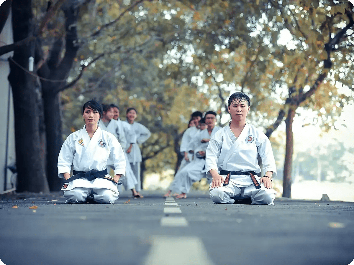 Two martial artists in white uniforms kneel on a tree-lined road, with a group of practitioners standing behind them, conveying focus and discipline.