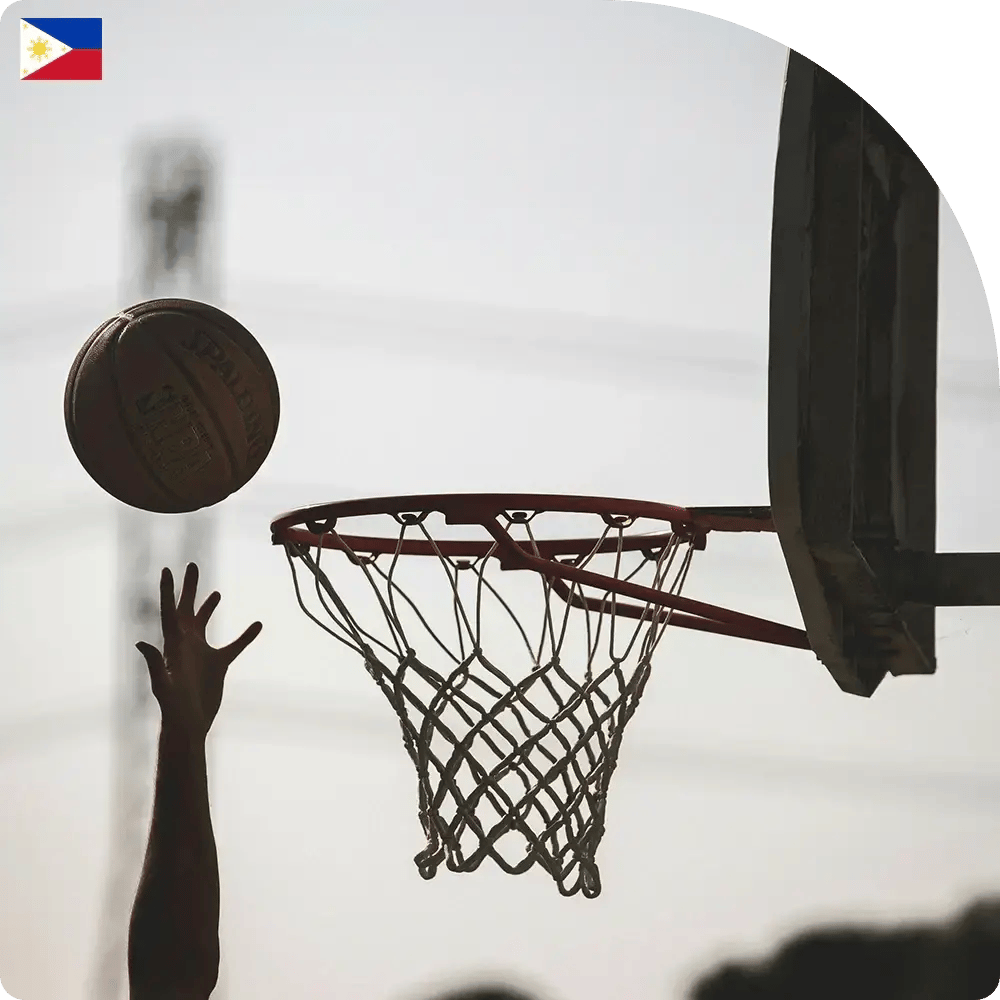 A silhouetted basketball player leaps towards the hoop, poised to shoot. The ball is mid-air against a blurred background of trees and a utility pole.