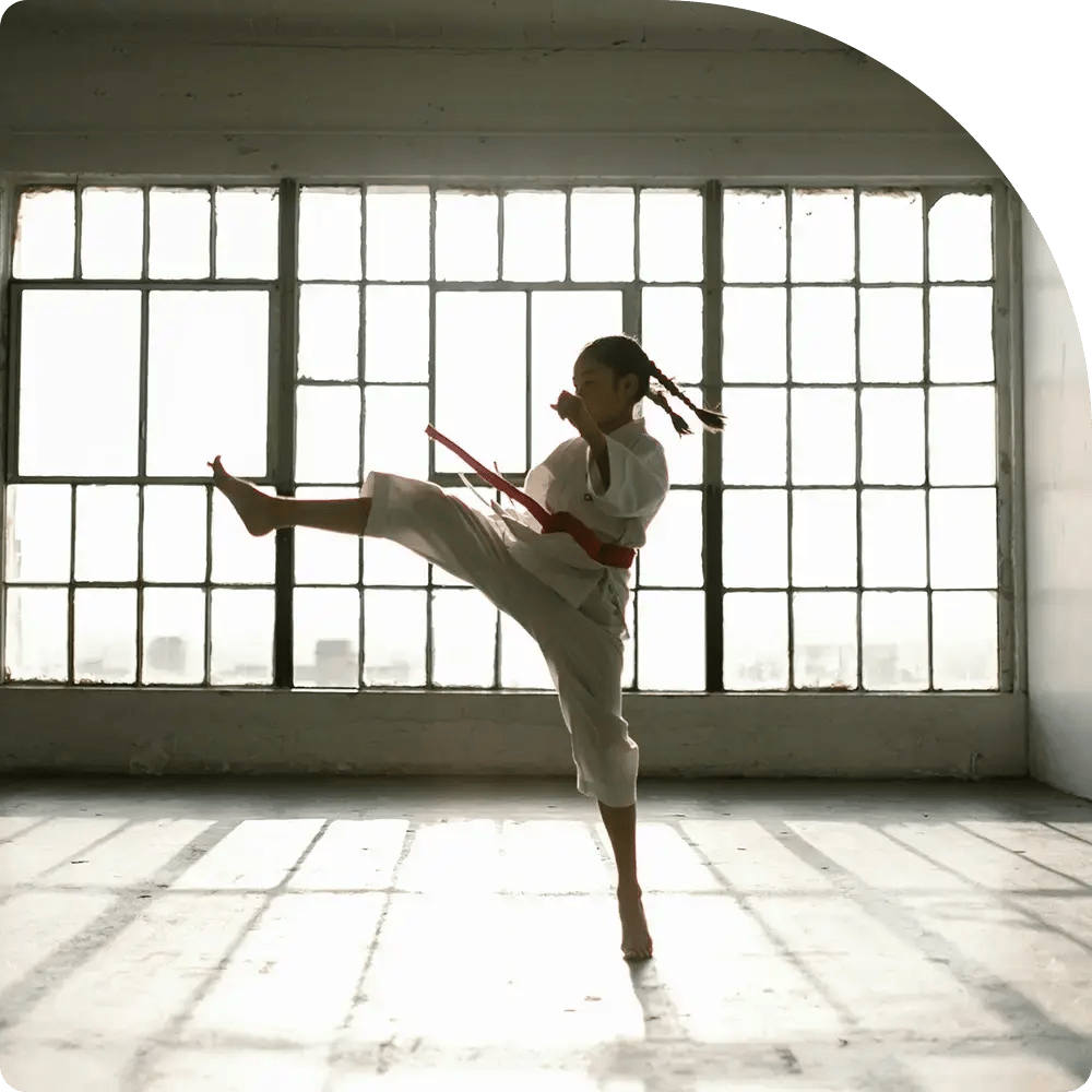 A young martial artist performs a high kick silhouetted against large industrial windows. She wears a white gi and red belt, exuding strength and focus.