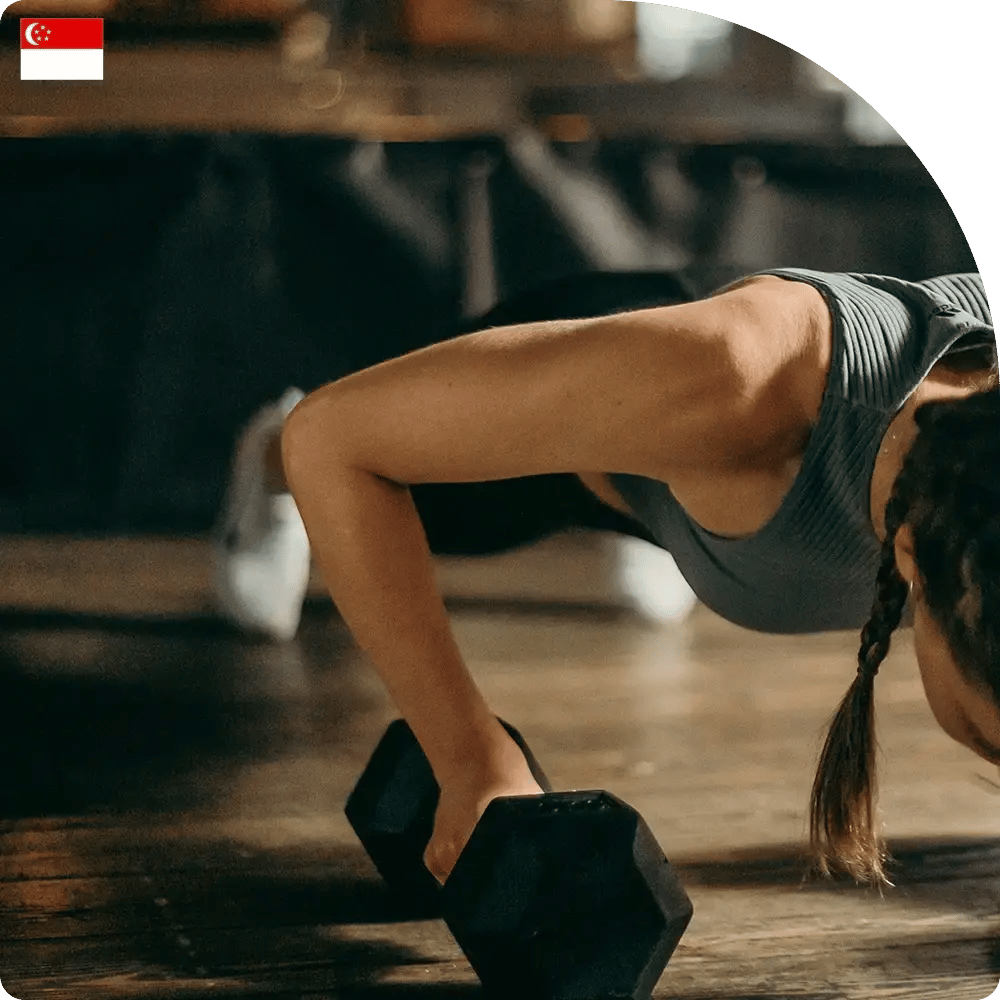 A person in a gray tank top performs push-ups using a dumbbell on a gym floor. The setting is dimly lit, conveying focus and intensity.
