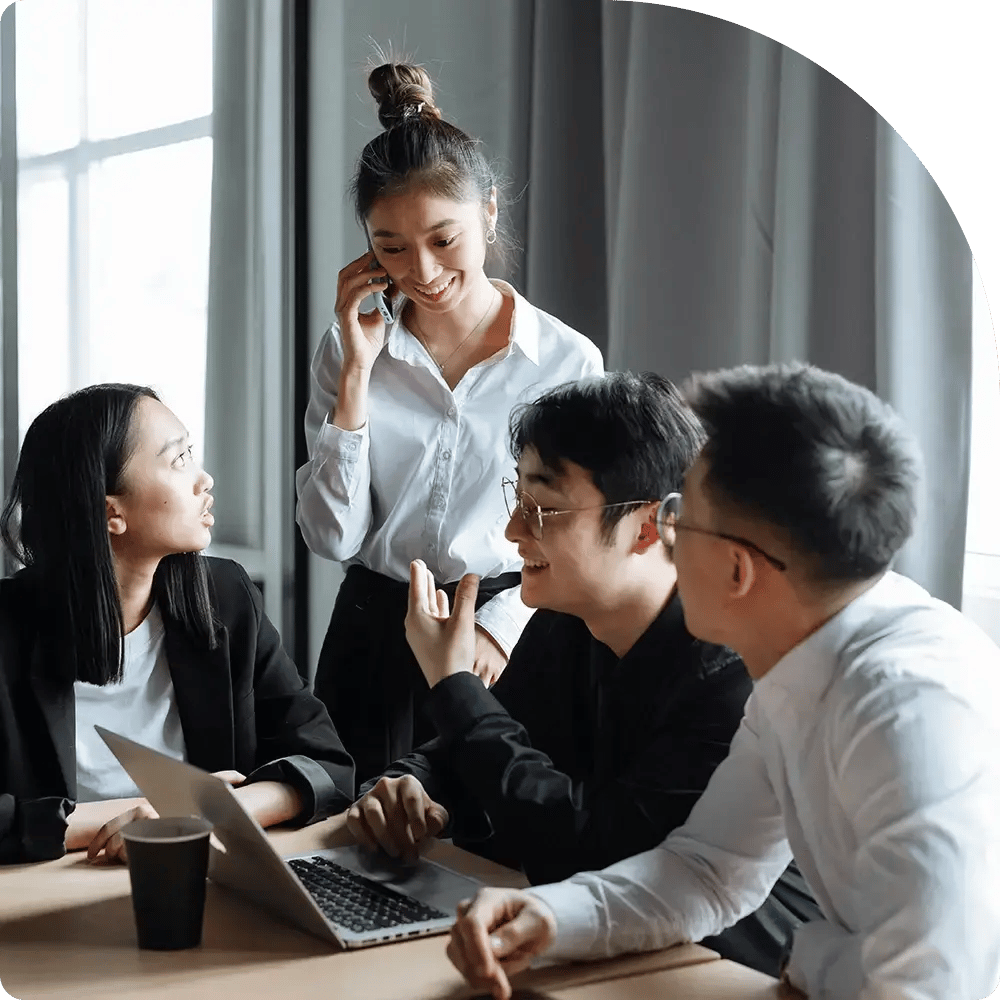 A group of four colleagues are having a lively discussion in an office. One person is on the phone, while others engage, smiling with a laptop nearby. The atmosphere is collaborative and positive.