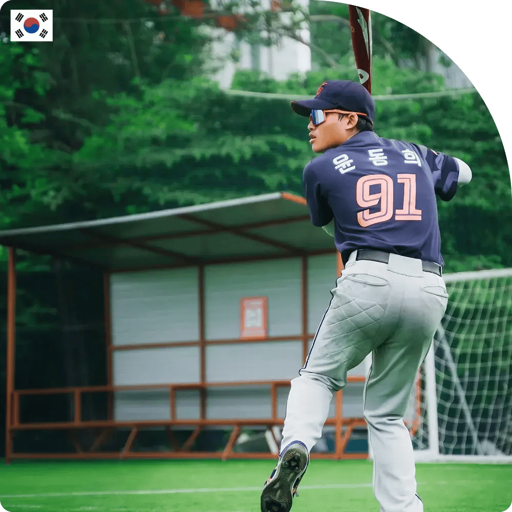 Baseball player in a blue jersey with number 91 swings a bat on a lush green field. A metal bench and goal net are visible in the background.