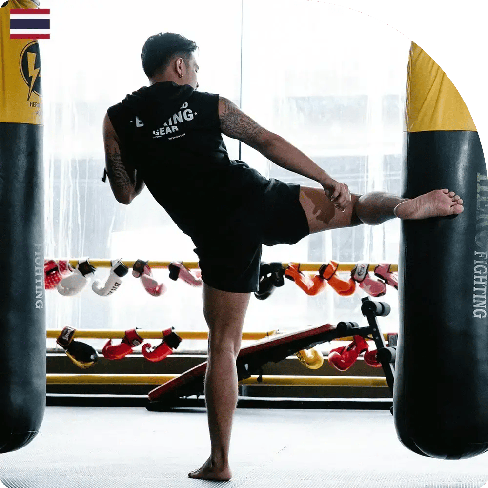 A person in a gym performs a side kick against a punching bag. They wear black athletic gear. Boxing gloves and benches are visible in the background.
