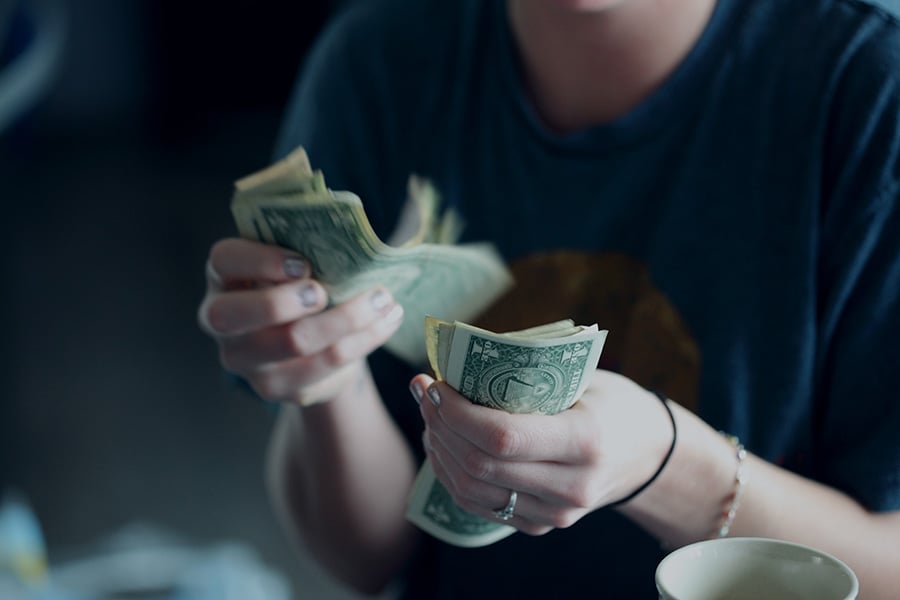 A person wearing a blue shirt counts a stack of U.S. dollar bills, focusing intently. 