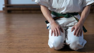 A person in a white karate gi with a green belt kneels on a wooden floor, hands resting on thighs, conveying focus and discipline in a dojo setting.