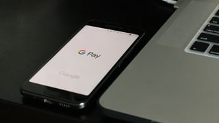 A smartphone displaying the Google Pay logo rests on a dark surface beside a silver laptop. The scene conveys a modern, digital payment setup.