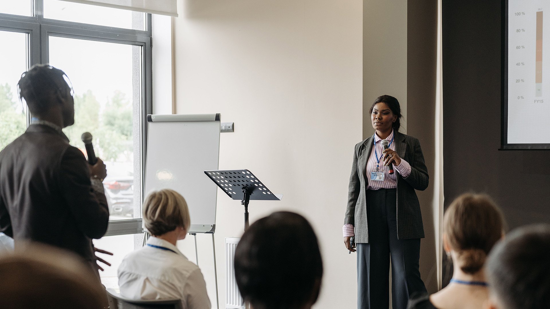 A woman presents at a conference, standing near a blank flip chart and projector screen. She looks focused, engaging an audience member holding a microphone.