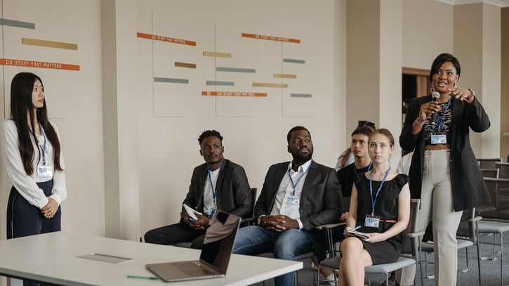 A diverse group of professionals in a meeting room. One woman stands, speaking confidently, while others sit attentively, fostering a collaborative atmosphere.
