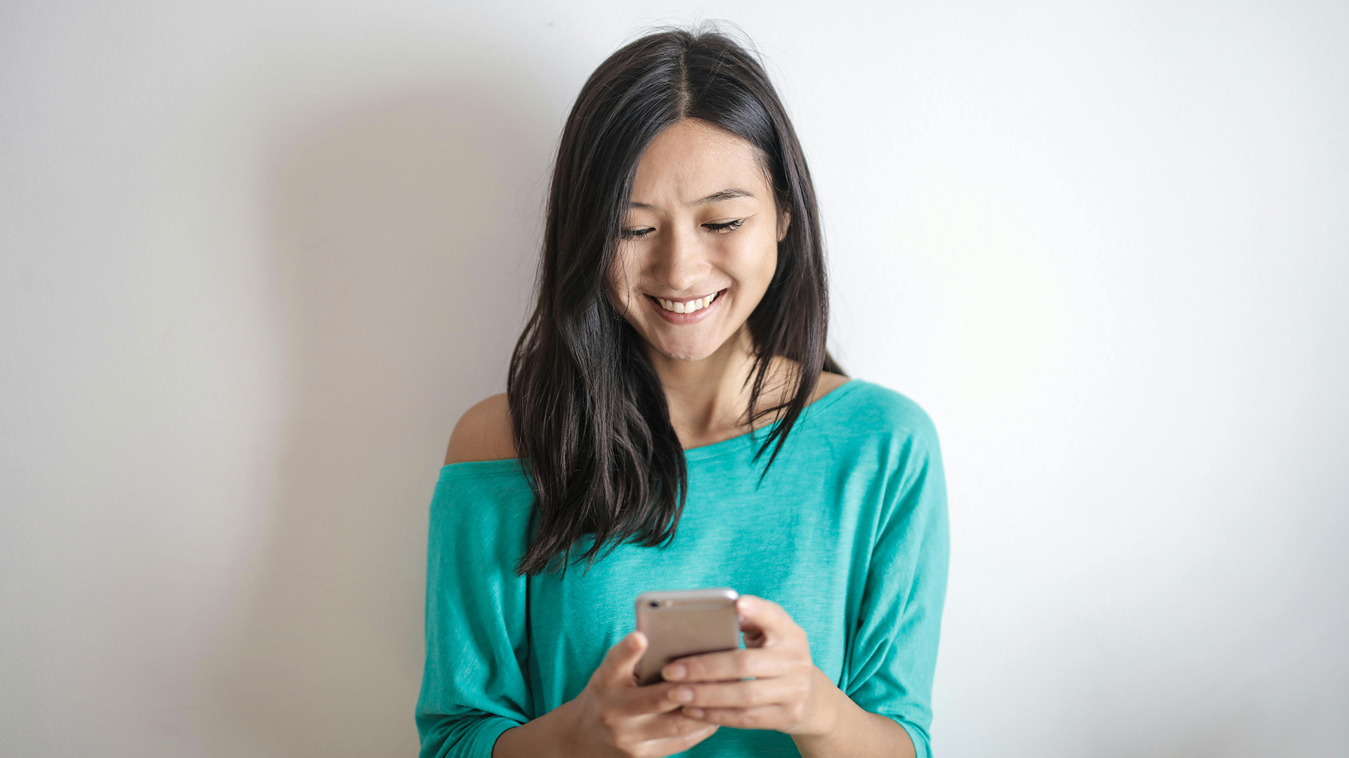 Smiling woman in a turquoise top looks at her smartphone against a plain white background, conveying a sense of happiness and engagement.