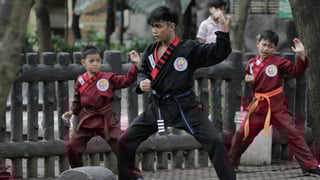 Three children practice martial arts outdoors, displaying focused expressions. The central child wears a black uniform, flanked by two in red.
