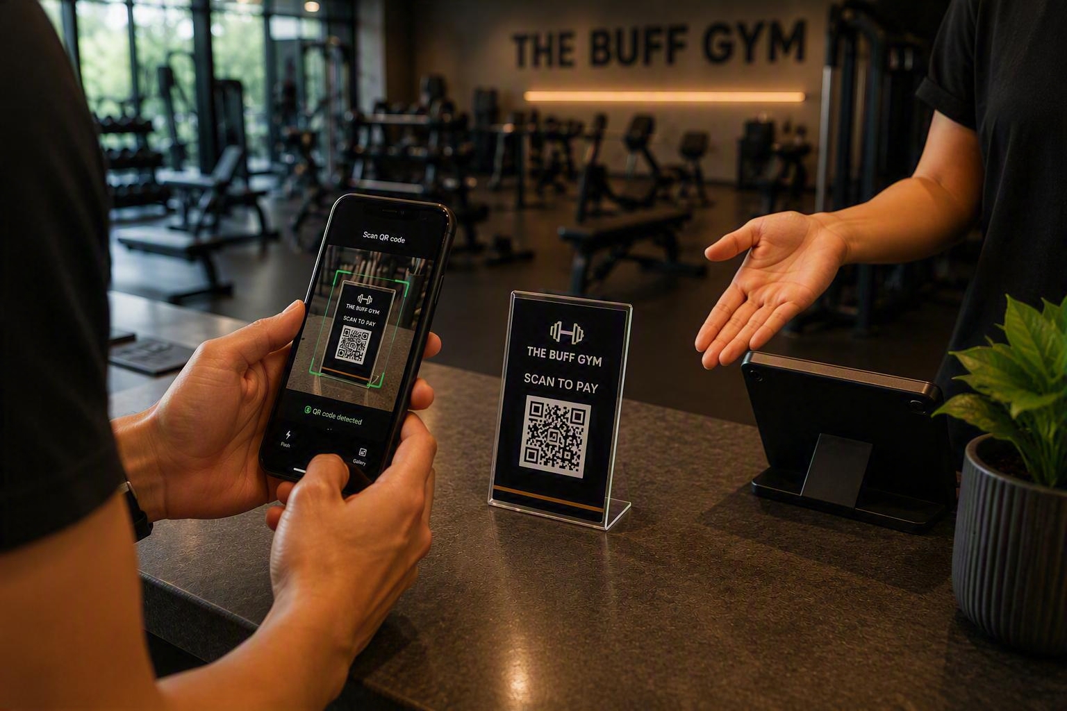 A gym goer scanning a QR code payment stand with a smartphone at The Buff Gym reception desk while staff direct them to complete a contactless payment.