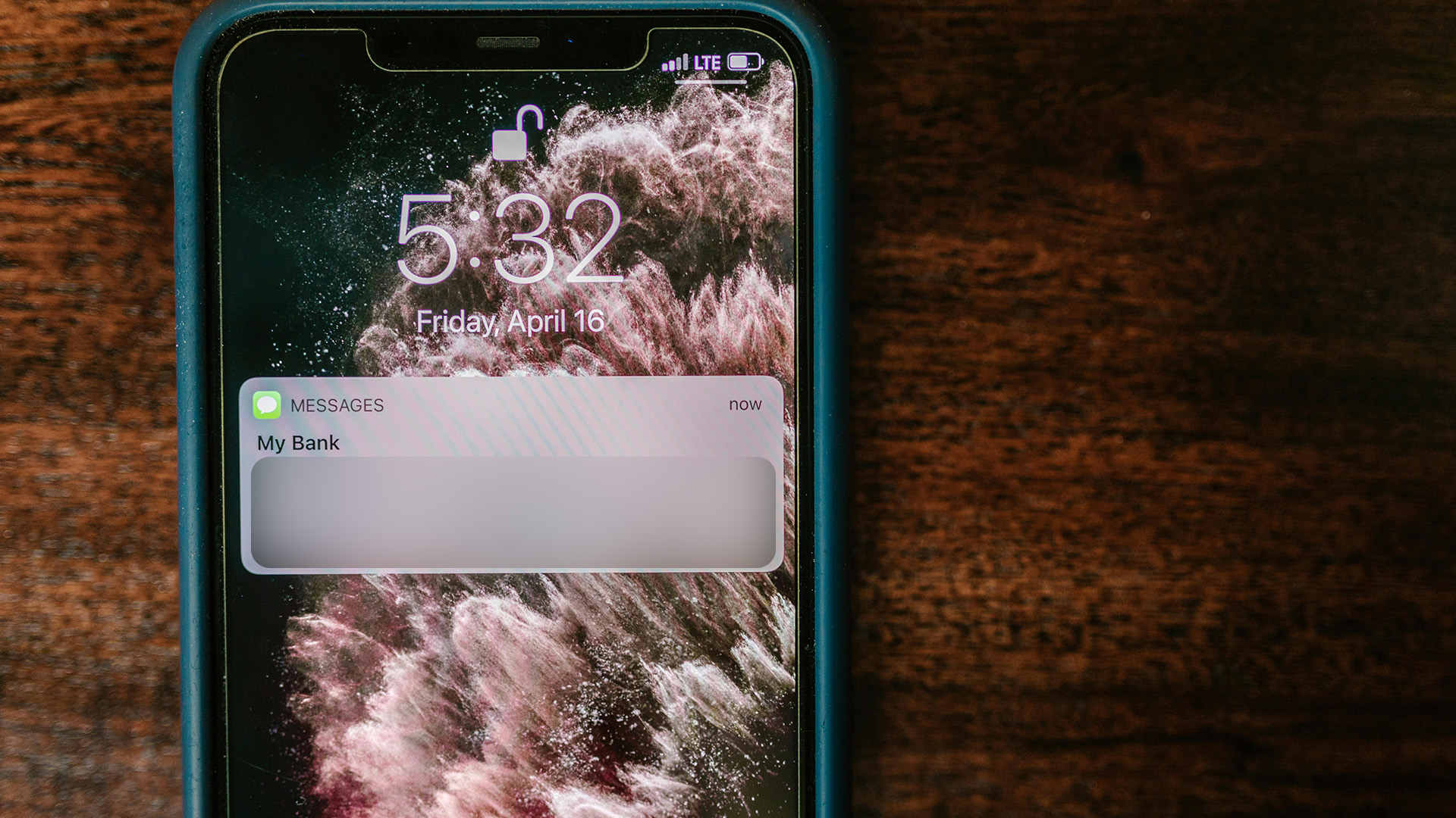 Smartphone with a lock screen reading 5:32 on April 16, displaying a notification from "My Bank." The phone rests on a dark wooden surface.