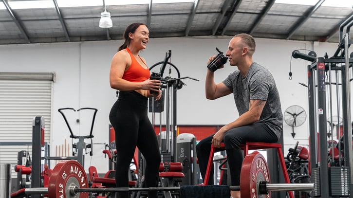 A woman in an orange sports bra and black leggings chats happily with a man in a gray shirt, both holding water bottles in a gym setting. They appear relaxed and engaged.