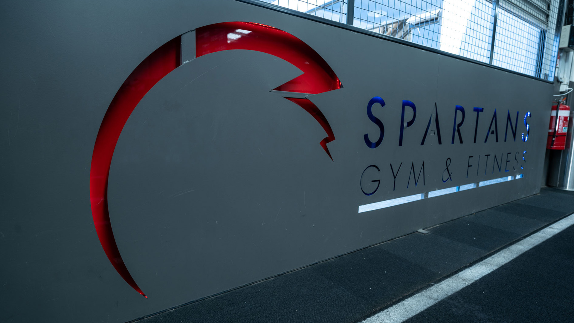 "Spartans Gym &amp; Fitness" sign with a stylized red Spartan helmet graphic. The setting is a modern gym with metal fencing.