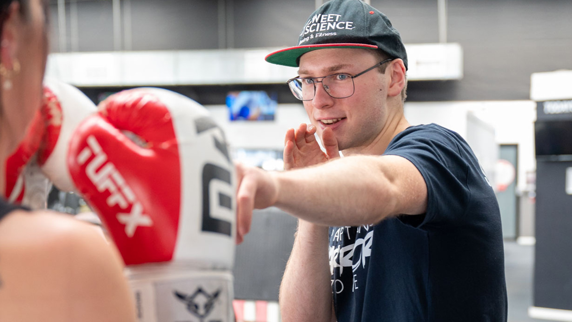 A boxing coach instructs a trainee in a gym, gesturing with a relaxed but focused expression. The trainee wears red boxing gloves, creating a dynamic training scene.