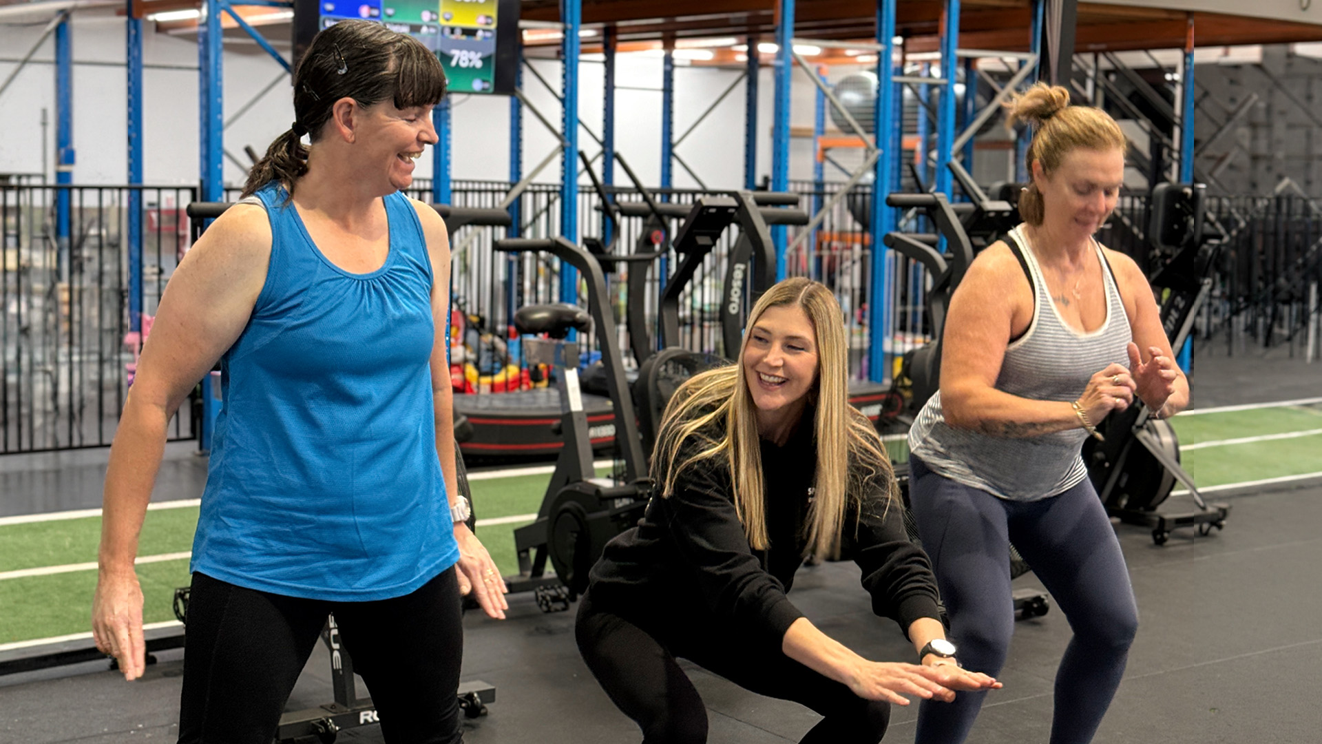 Three women exercising together in a gym, one squatting while others smile. The atmosphere is energetic and friendly, with gym equipment in the background.